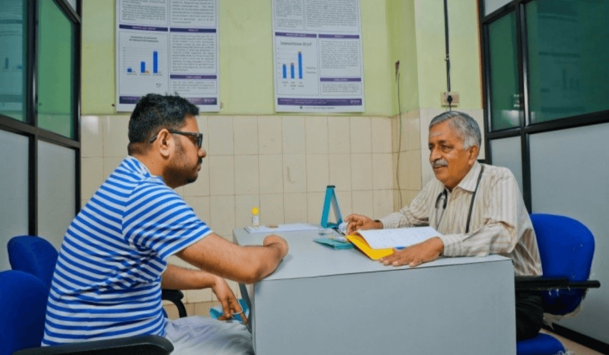 A man wearing a stethoscope around his neck talks across a desk to a patient