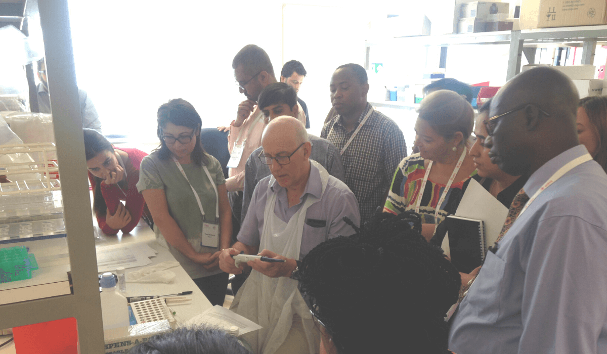In a medical lab, a group of people crowd around a man giving a demonstration