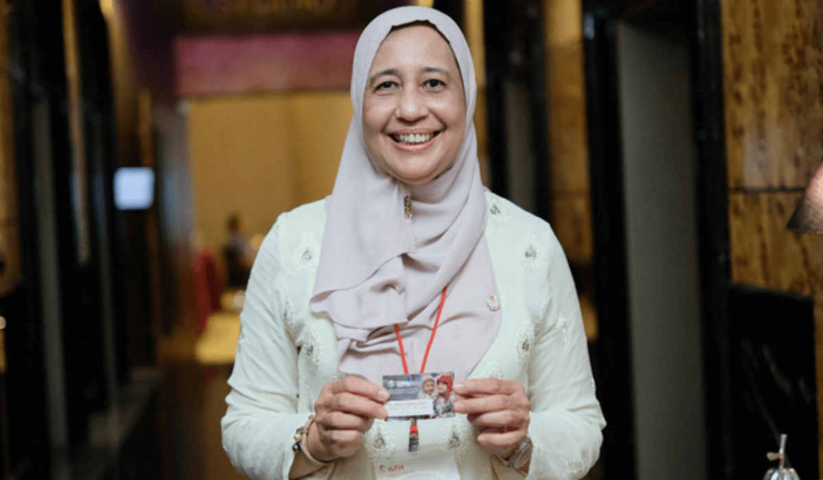 A woman in a beige headscraf smiles at the camera, holding a small flyer
