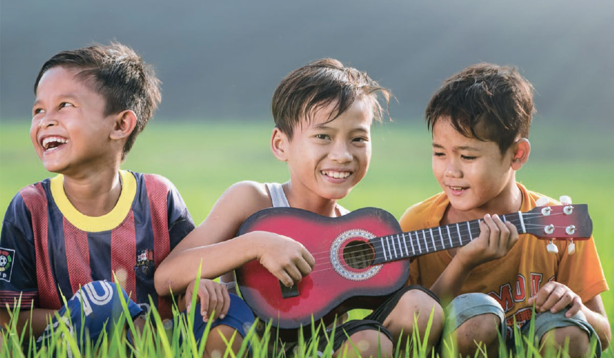 Tres niños sentados juntos y sonrientes; uno de ellos toca un instrumento.